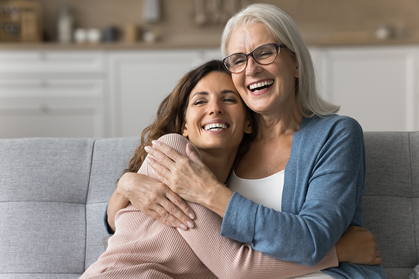 Cheerful attractive senior mom and happy adult daughter hugging at home with love, care, resting on couch together, smiling, laughing, talking, enjoying family relationship, bonding, motherhood