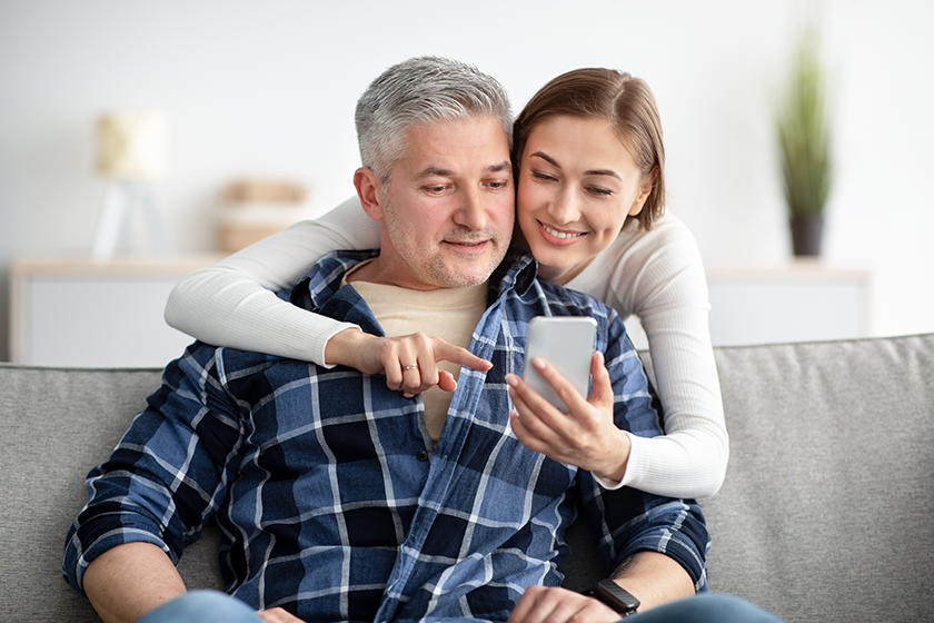 Cheerful mature couple using cellphone while resting on couch in living room